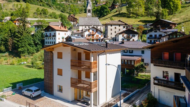 A modern building with a balcony, a parking area, and a view of a town with a church and houses. - Casa Carducci (Alleghe)