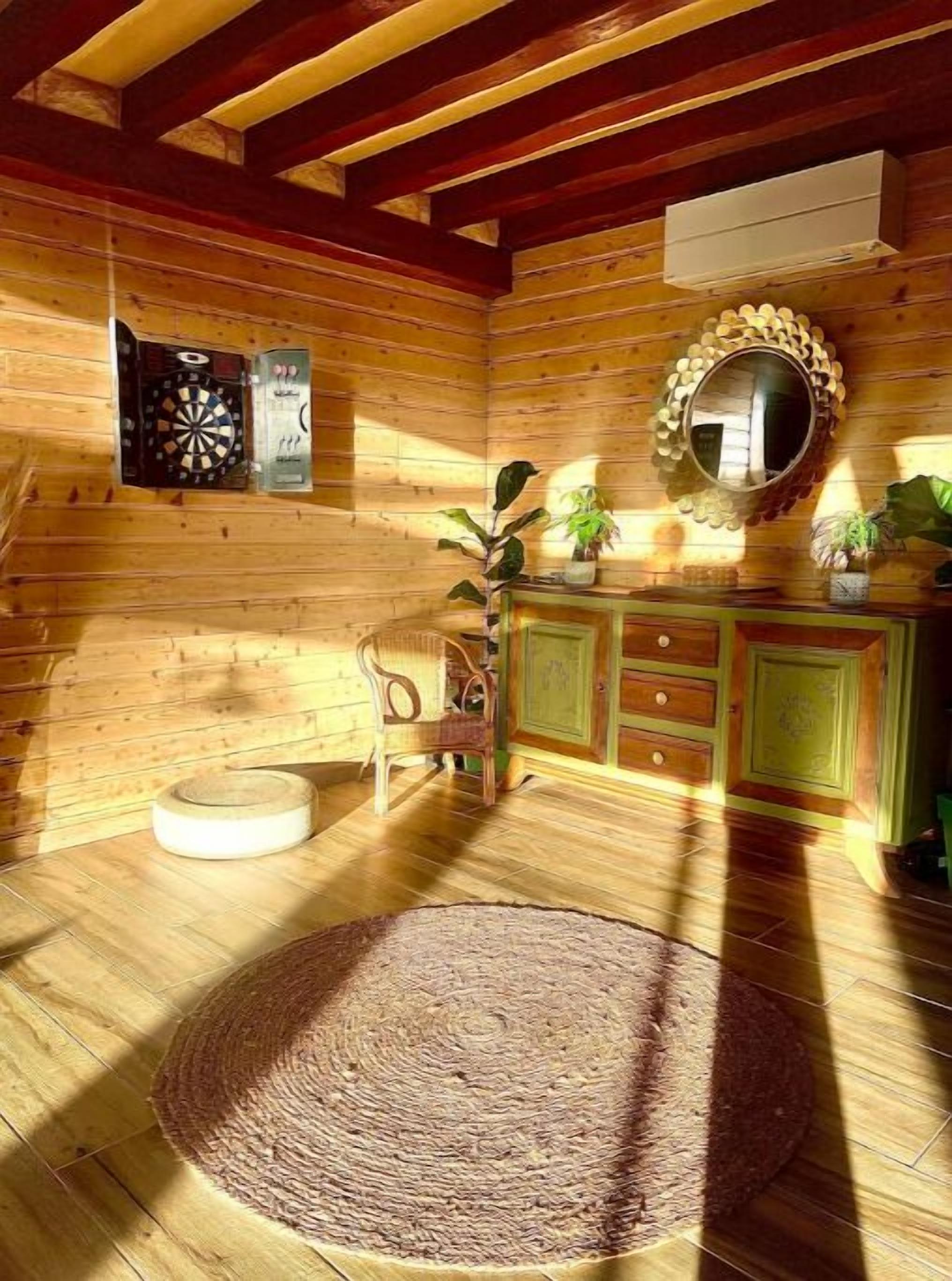 L’intérieur d’un chalet en bois, avec un jeu de fléchettes, un miroir, une chaise, une petite table et un tapis rond.