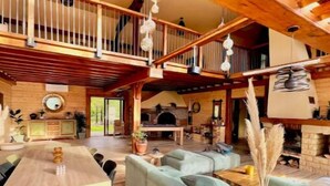 A spacious living area with a high ceiling, wooden beams, a dining table, sofas, and a fireplace. - Chalet Acacia au Calme 1h30 de Paris (La Selle-en-Hermoy)