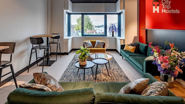 Een moderne hostelkamer met een groene bank, een zithoek met een tafel en stoelen, een groot raam met uitzicht op de stad, en een rode muur met het logo van het hostel.