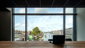Een kamer met een groot raam dat uitkijkt op een gracht en gebouwen, een houten tafel met een laptop en muis, en een stoel.