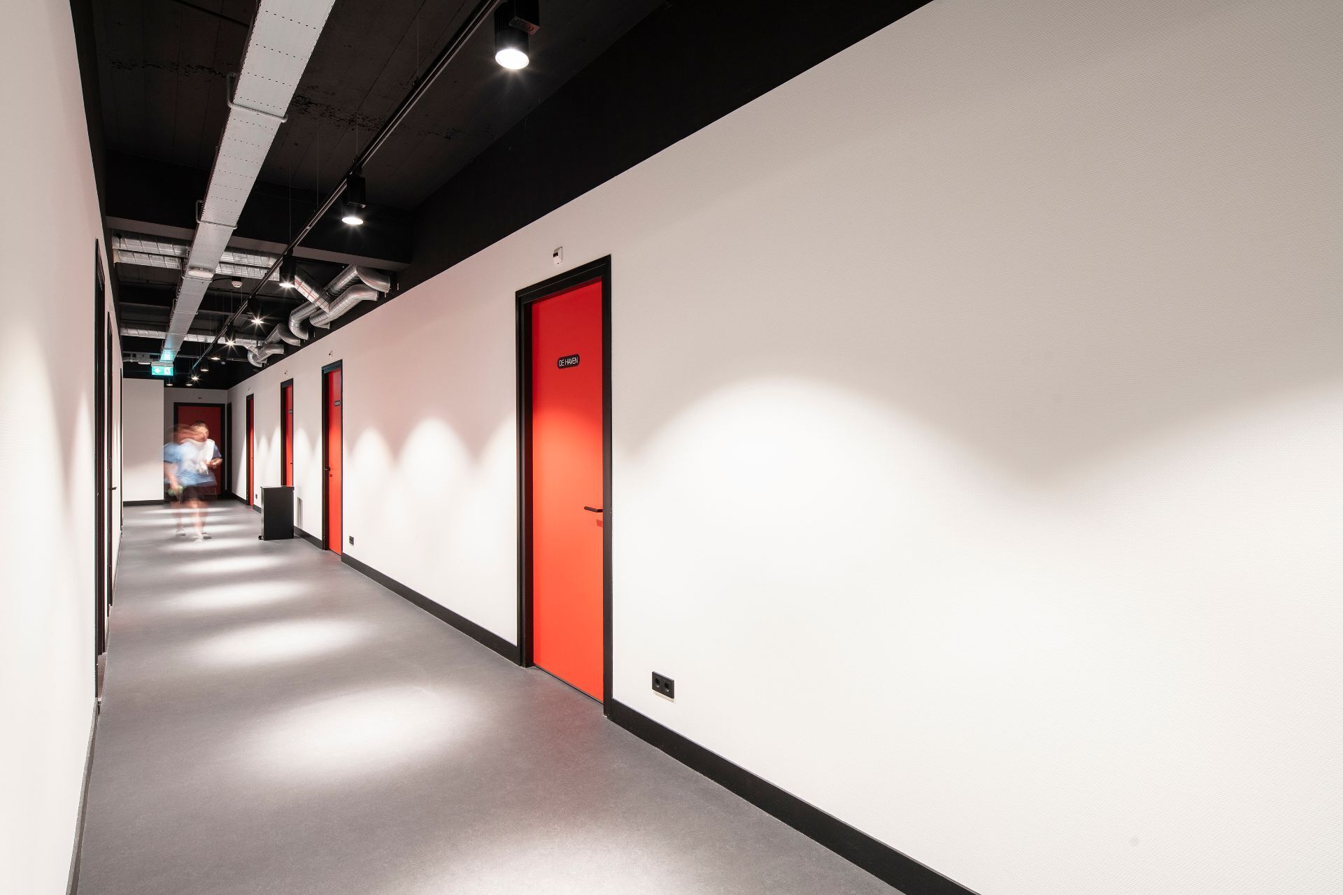 A long, narrow hallway with white walls and red doors, featuring a modern industrial ceiling and lighting.