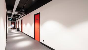 A long, narrow hallway with white walls and red doors, featuring a modern industrial ceiling and lighting.