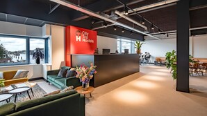 Een moderne hotellobby met een receptiebalie, een zithoek en grote ramen met uitzicht op een stadsbeeld.