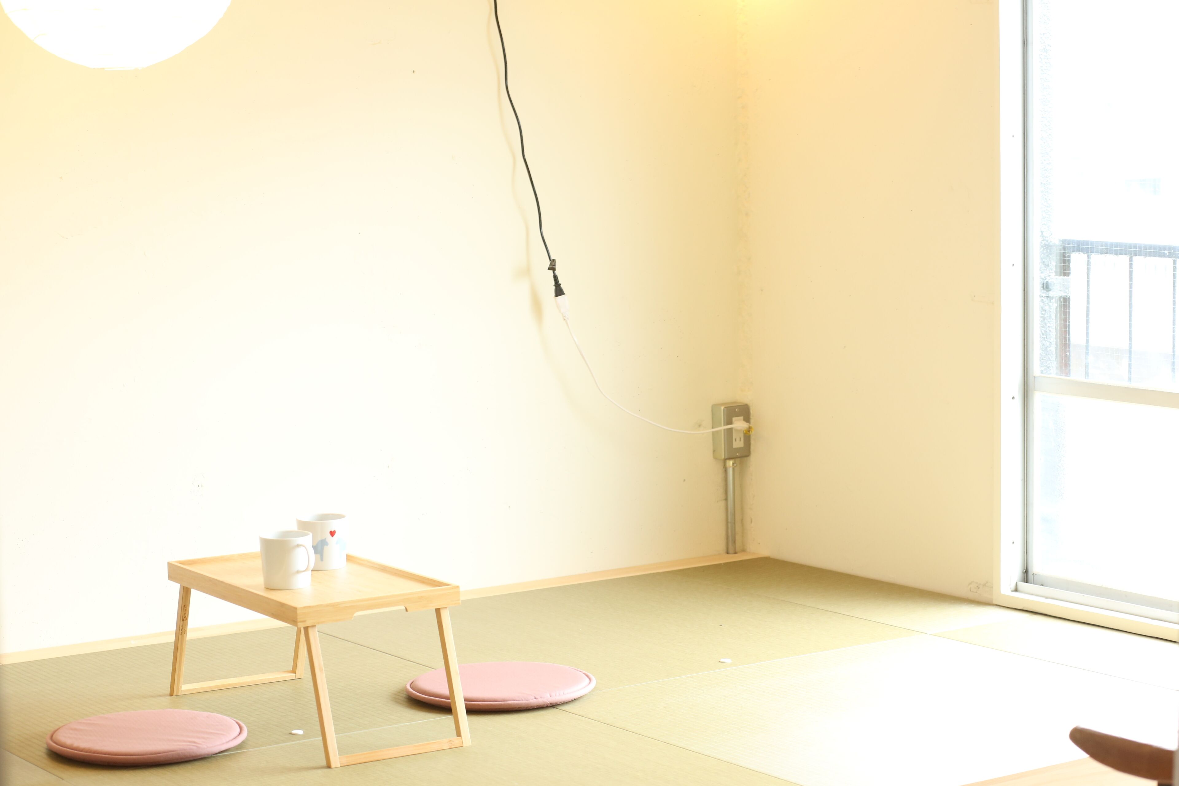 Una habitación minimalista con suelo de tatami, una mesa de madera con dos cojines rosados y un ventanal con vistas al exterior.