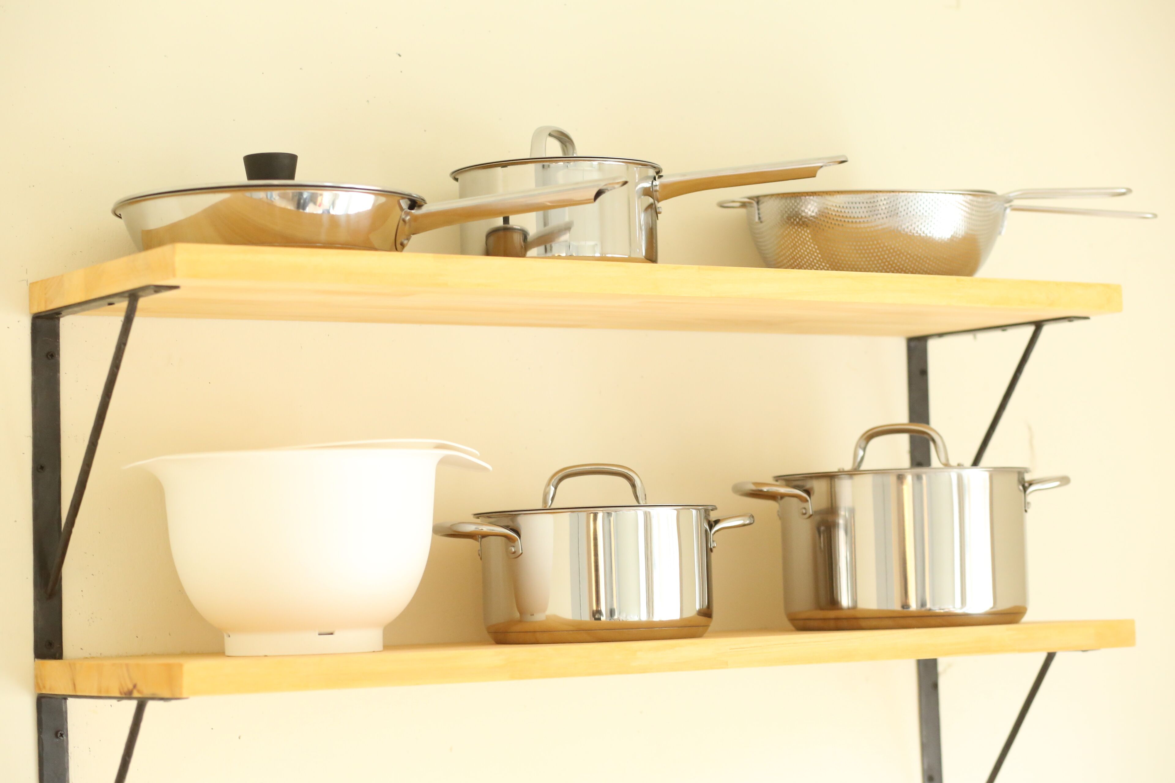 A kitchen shelf with various stainless steel pots and a white bowl.