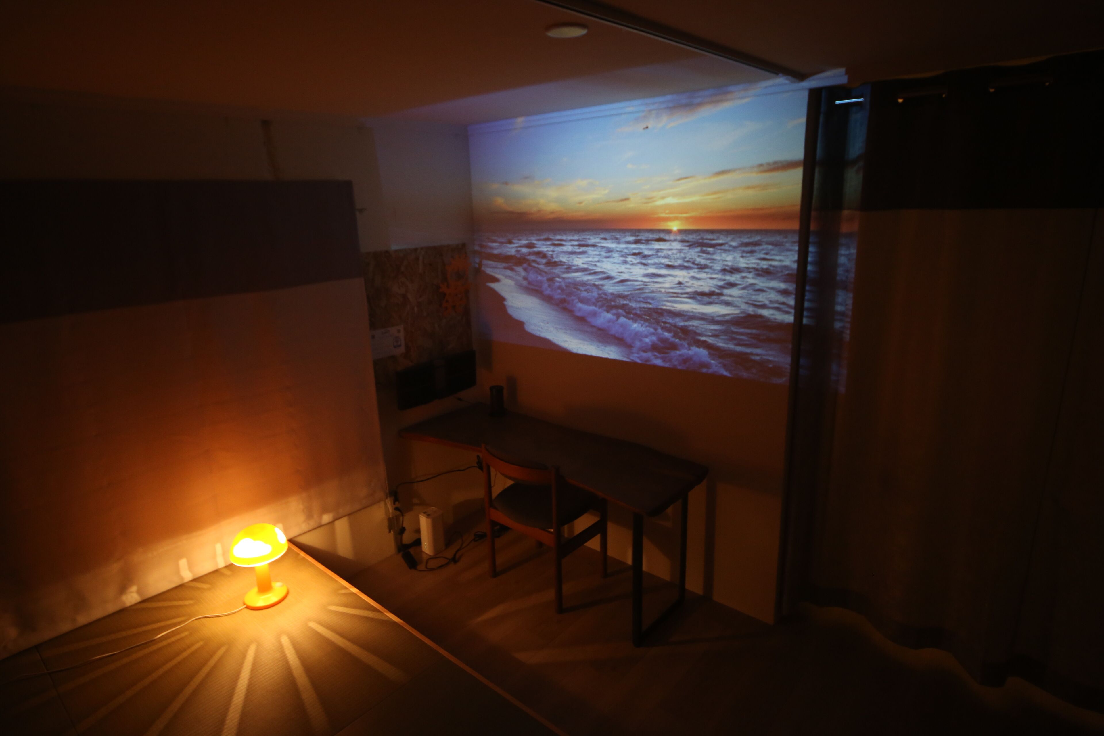 A dimly lit room with a wall-mounted projector displaying a sunset over the ocean. A desk with a chair is positioned under the projection.