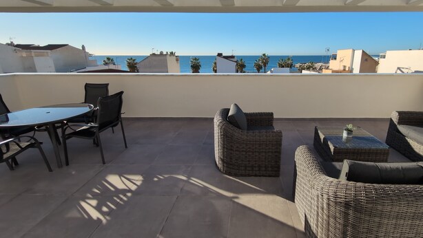 A terrace with wicker furniture, a table, and chairs, overlooking a coastal view with buildings and palm trees. - Penthouse El Carmelo New, Modernly Furnished with Huge Terrace < 50m from Beach (Caleta de Vélez)