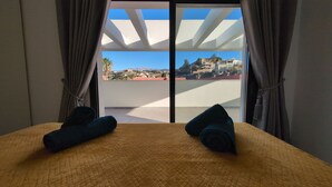 A room with a bed, two rolled towels, and a view of a hillside with buildings. - Penthouse El Carmelo New, Modernly Furnished with Huge Terrace < 50m from Beach (Caleta de Vélez)