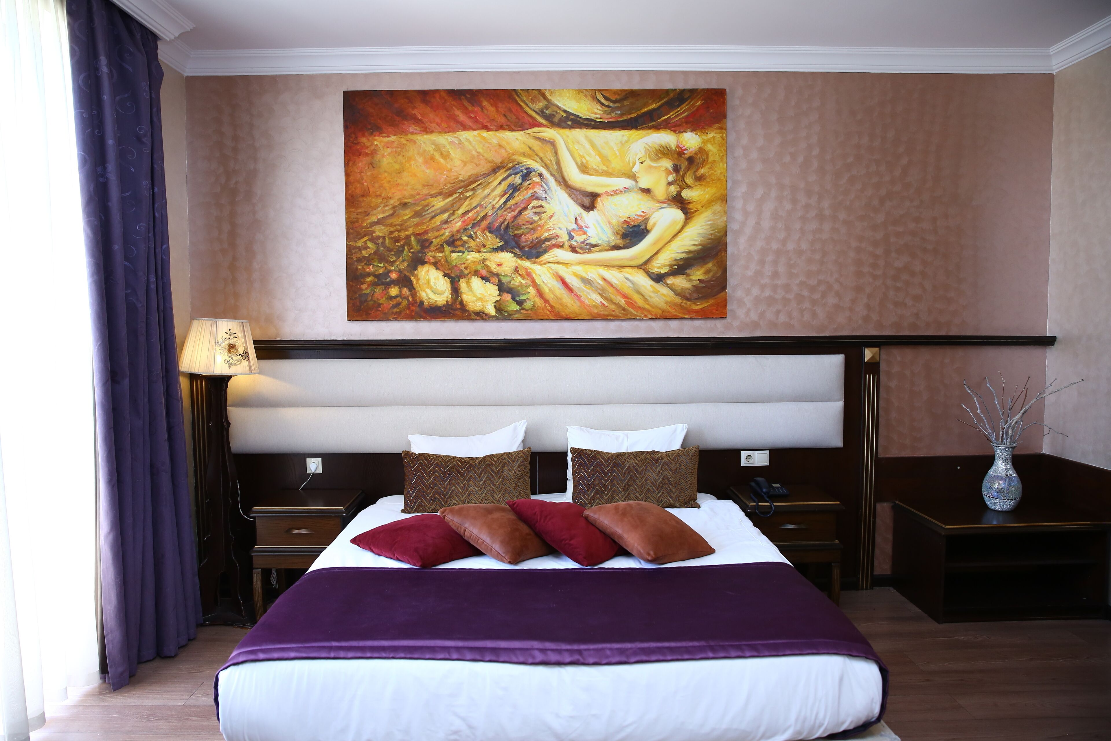 A hotel room with a large bed, a painting above the bed, a nightstand, a lamp, and a vase on a side table.