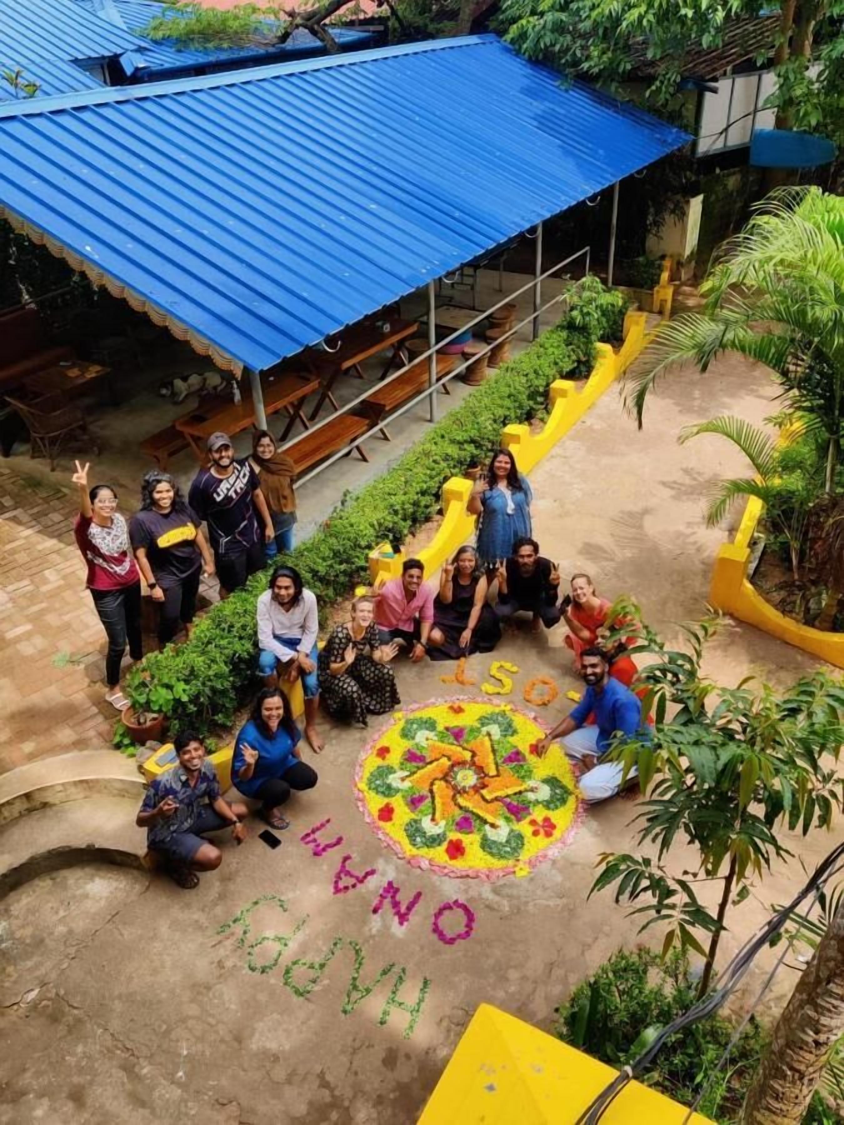 A group of people gathered around a colorful rangoli on a courtyard with a blue-roofed structure in the background.