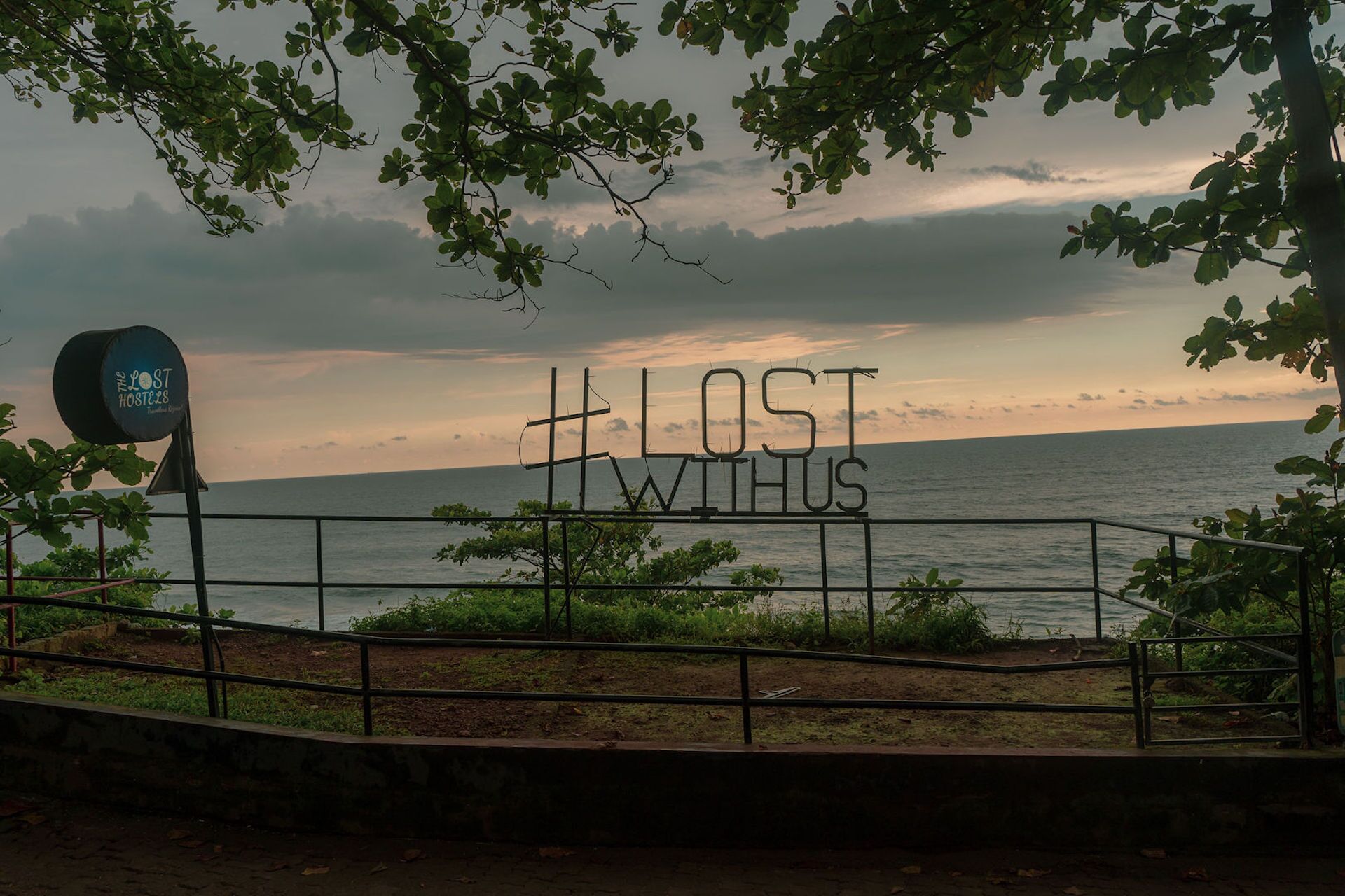 A sign with the text "LOST WITH US" against a backdrop of a sunset over the sea, with a metal fence and greenery in the foreground.