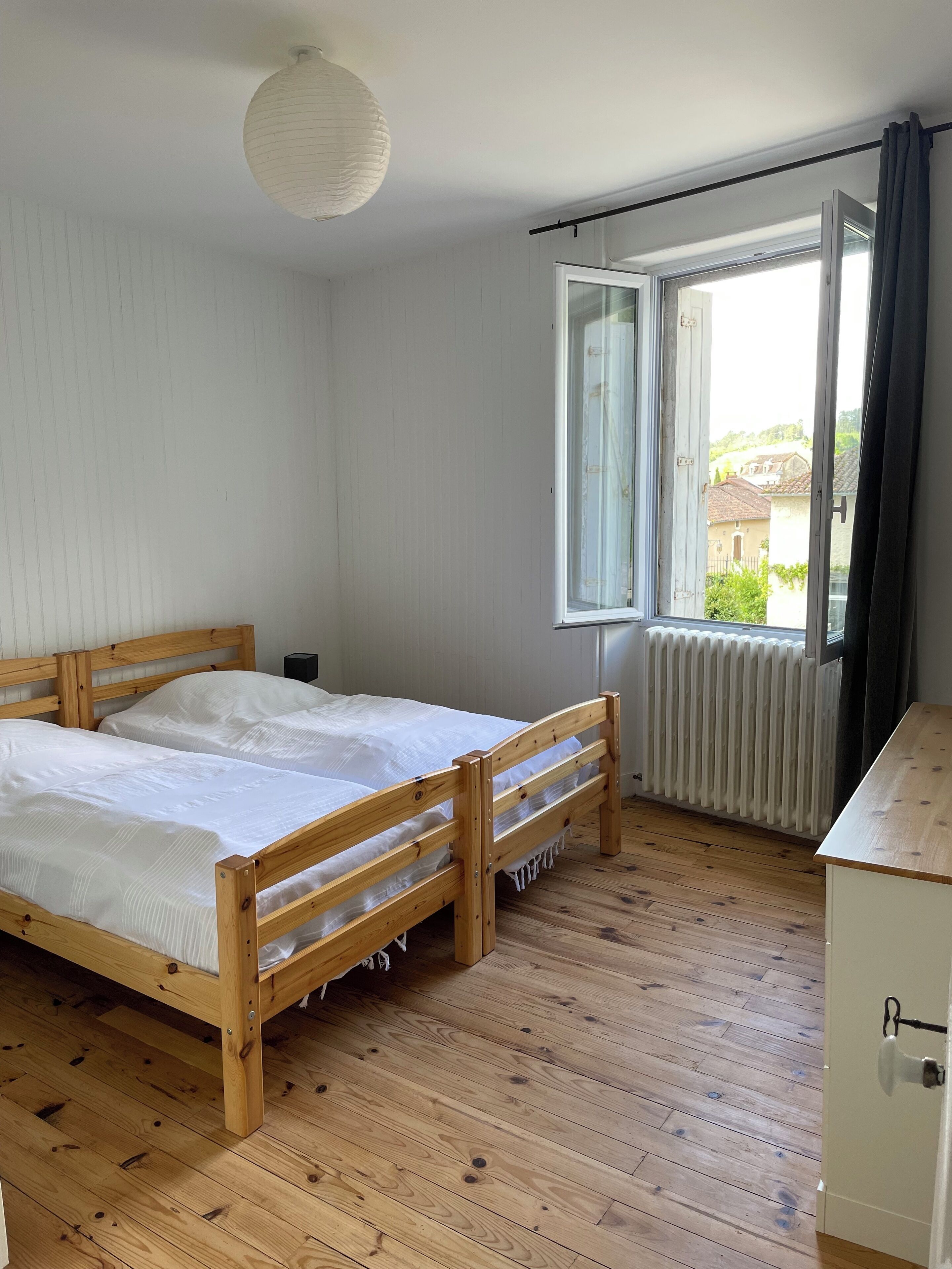 Ein Schlafzimmer mit einem Holzbett, einem mit Vorhängen versehenen Fenster, einer Heizung und Blick ins Freie.