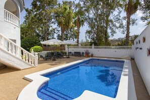Une piscine dotée d’une terrasse blanche, entourée de chaises longues et de parasols, et un bâtiment blanc avec un balcon.