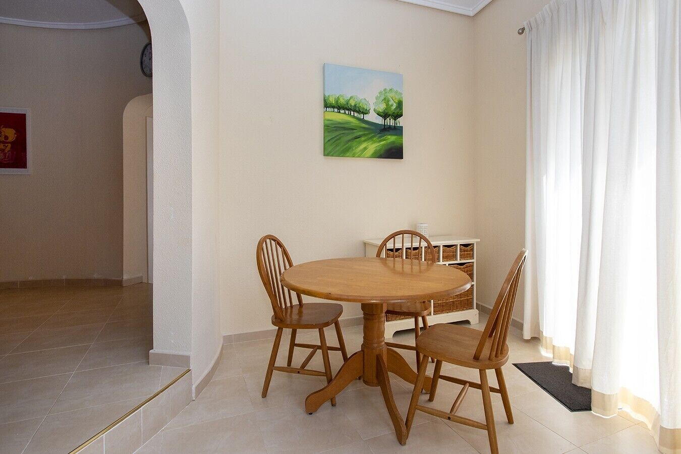 A dining area with a round wooden table and four chairs, a white cabinet with baskets, and a painting of trees on the wall.