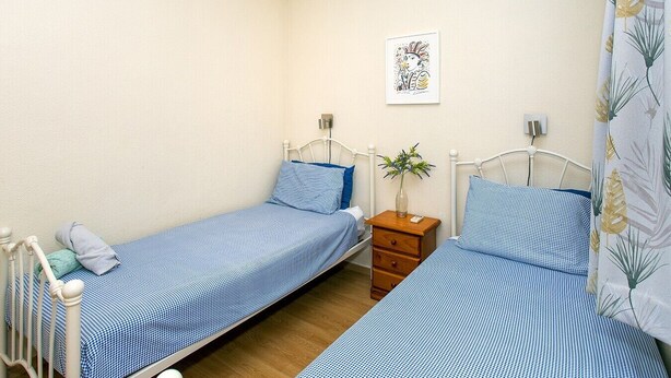 Un alojamiento pequeño y de una sola habitación con dos camas individuales, una mesita de noche con un jarrón y una lámpara fijada a la pared.
