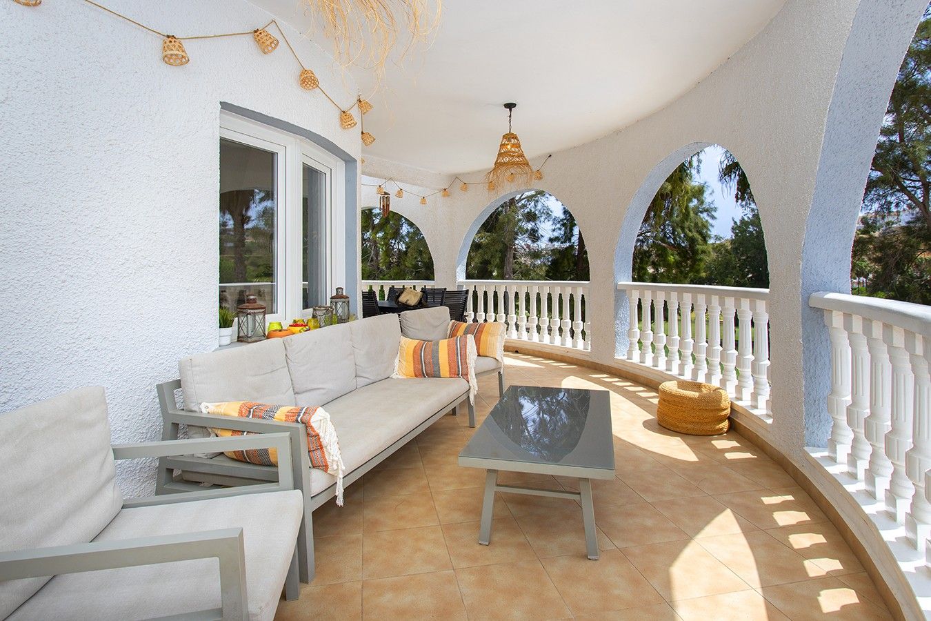 A covered outdoor seating area with a sofa, chairs, and a table, surrounded by white railings and a pergola with string lights.