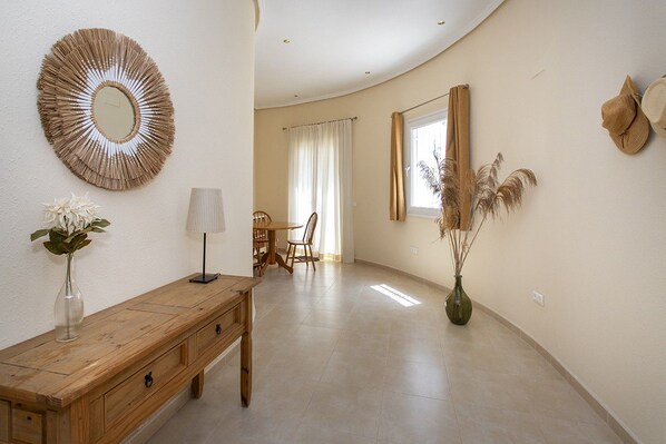 A hallway with a wooden bench, a mirror, a vase with flowers, a lamp, and a window with curtains.