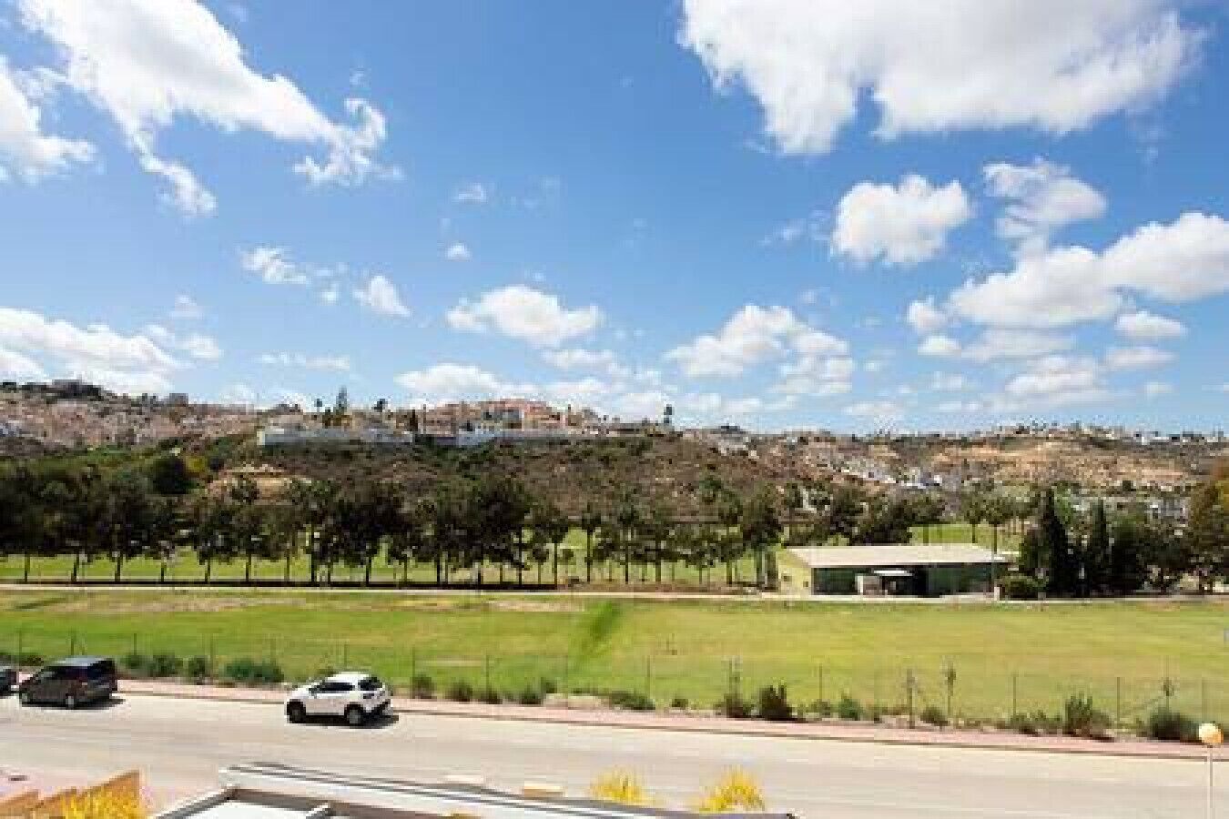 Carretera amb vehicles, una zona verda i un edifici amb teulada plana al fons.