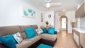 A living room with a sofa, teal cushions, a ceiling fan, and a kitchen area with a counter and cabinets.