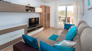 A modern living room with a flat-screen TV, wooden shelving, a sofa with blue cushions, and a sliding glass door leading to an outdoor area.