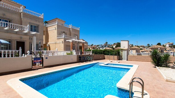 A swimming pool with a clear blue water, surrounded by a tiled deck, and adjacent to a building with balconies and a view of the surrounding area.