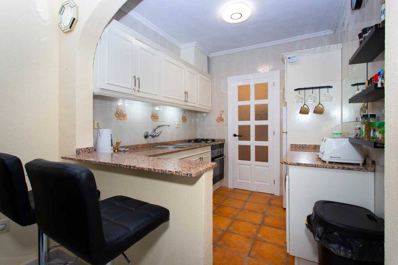 Cocina compacta con gabinetes blancos, encimera de granito, horno empotrado y una pequeña zona de comedor con una silla negra.