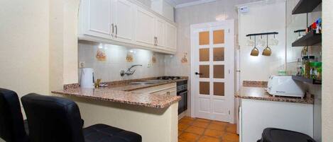 Cocina compacta con gabinetes blancos, encimera de granito, horno empotrado y una pequeña zona de comedor con una silla negra.