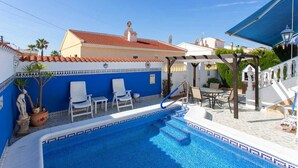 Piscina com borda azulejada em azul, uma cadeira branca, um guarda-sol branco e uma pérgola com área de estar ao ar livre.