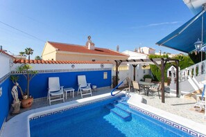 Une piscine dont le bord est recouvert de carreaux bleus, une chaise blanche, un parasol blanc et une pergola avec des sièges d’extérieur.