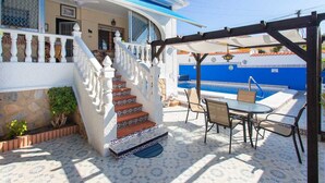 Uma casa com fachada branca, detalhes em azul, um pátio com azulejos, uma escadaria e uma área de jantar com mobiliário para área externa.