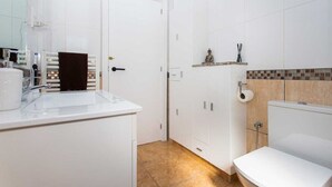 Casa de banho com lavatório branco, sanita e área de chuveiro. Uma porta dá para outro espaço, e uma pequena prateleira exibe produtos de higiene pessoal.