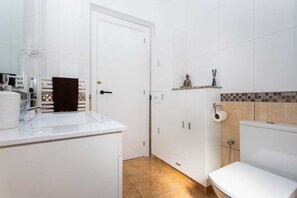 Une salle de bain avec un lavabo blanc, des toilettes et un espace douche. Une porte donne accès à une autre pièce, et une petite étagère contient des articles de toilette.
