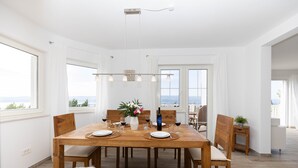 Een eethoek met een houten tafel voor zes personen, witte stoelen en uitzicht op zee door grote ramen.