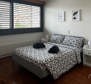 Ein Schlafzimmer mit einem Bett, einem Nachttisch, zwei gerahmten Bildern an der Wand und Jalousien am Fenster.