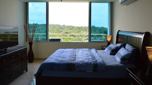 Une chambre à coucher comprenant un lit, un téléviseur, un ventilateur de plafond et une grande fenêtre donnant sur un paysage verdoyant.