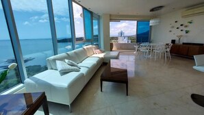 A modern living room with a white sofa, a coffee table, and a dining area with white chairs. Large windows offer a view of the ocean and a cityscape. - Luxury Paradise at Playa Bonita (Panama City)