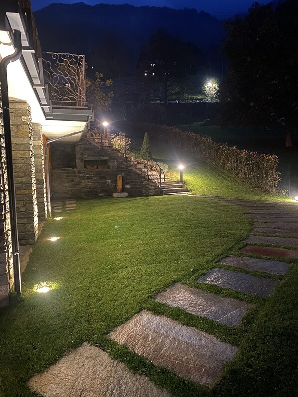 A well-lit stone pathway leading to a stone building with a modern design, surrounded by a grassy area and a hilly landscape in the background. - Bed and Breakfast Soffio di Vento (Perloz)