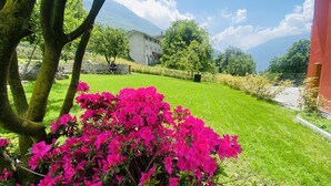 A well-maintained lawn with vibrant pink flowers in the foreground, a stone wall, and a building with a sloped roof in the background. - Bed and Breakfast Soffio di Vento (Perloz)