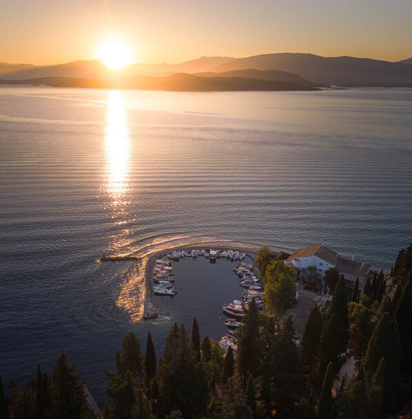 A marina with boats docked in a semi-circle, surrounded by trees and overlooking a calm sea at sunset.
