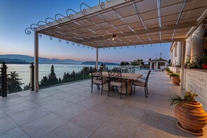 A covered outdoor patio with a table and chairs, overlooking a body of water and mountains.