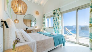 Un dormitorio con cama, escritorio, silla, ventana grande con cortinas y vista al mar.