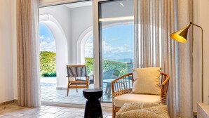 Ein Zimmer mit einer großen Schiebeglastür, die auf eine Terrasse im Freien führt, einem Korbssessel, einem runden schwarzen Beistelltisch und einer Stehlampe.