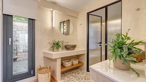 A modern bathroom with a stone wall, a round mirror, a sink, a potted plant, and a sliding door. - Agios Stefanos Bay - Villa Maria (Agios Stefanos Kassiopi)
