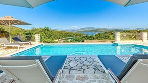 A pool with lounge chairs and umbrellas, surrounded by stone tiles, with a view of mountains and a body of water. - Agios Stefanos Bay - Villa Maria (Agios Stefanos Kassiopi)