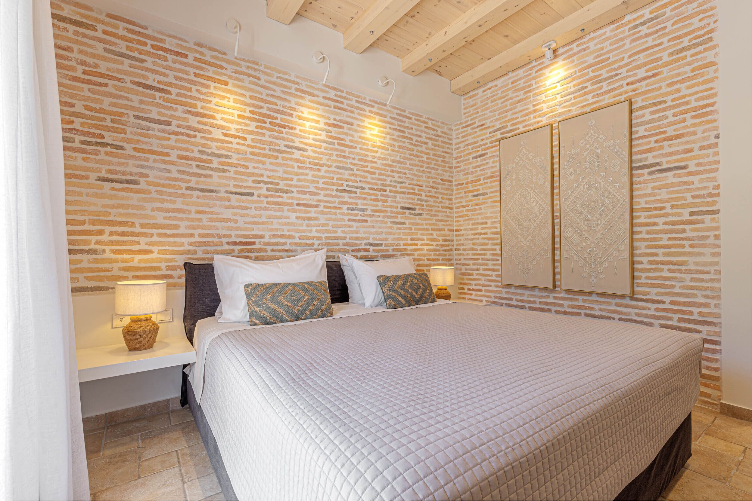 Ein Schlafzimmer mit einer Wand im Ziegelstein-Muster, einem großen Bett mit gesteppter Bettdecke, zwei Nachttischlampen und einem Wandbild.