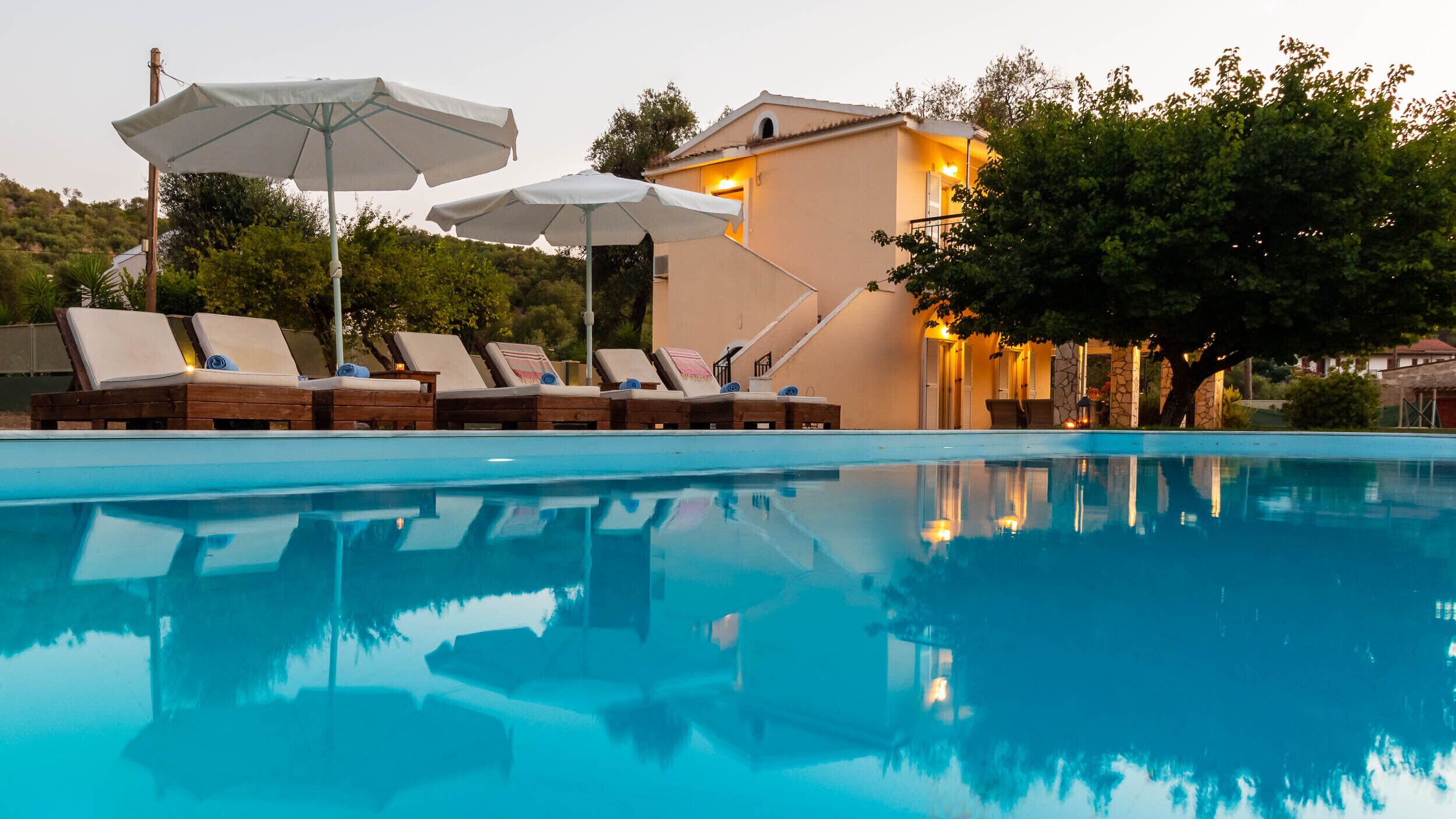 A swimming pool with lounge chairs and umbrellas, a building with a stone facade, and trees in the background.