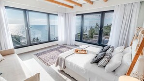A bedroom with a large bed, two windows with curtains, a view of the sea, and a wooden ceiling.