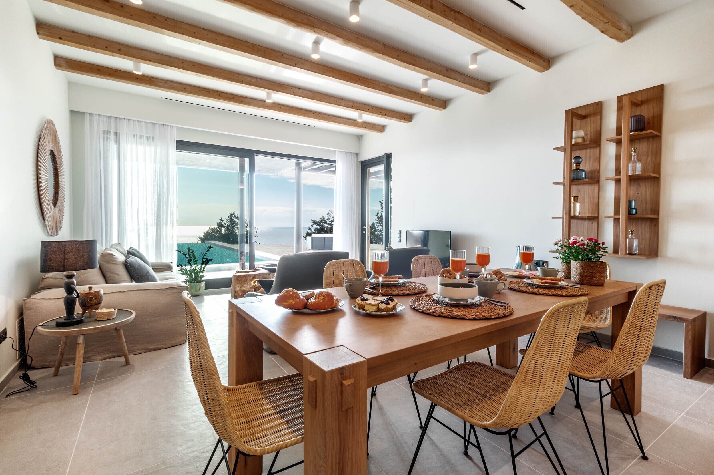 Un salon moderne avec une table à manger en bois, des chaises en osier, un canapé, un téléviseur et une grande fenêtre donnant sur un beau paysage.