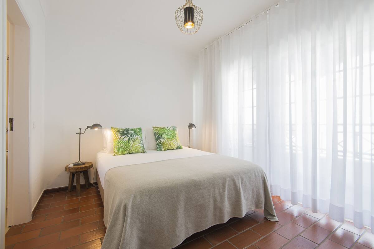 Une chambre à coucher avec un lit, deux lampes de chevet, une table de chevet et une fenêtre avec des rideaux légers.